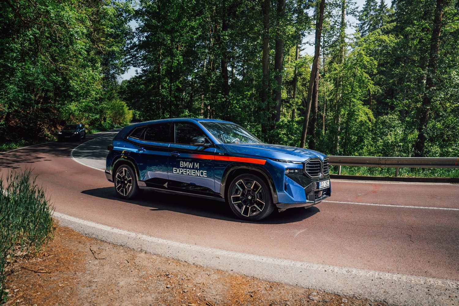 Blue BMW XM with M Experience branding driving on a scenic forest road on a sunny day.