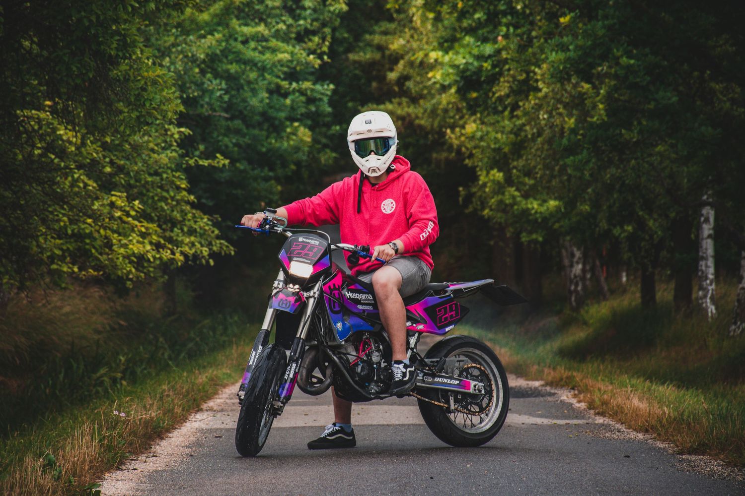Rider in red hoodie sits on purple-pink dirt bike on forest road wearing white helmet.
