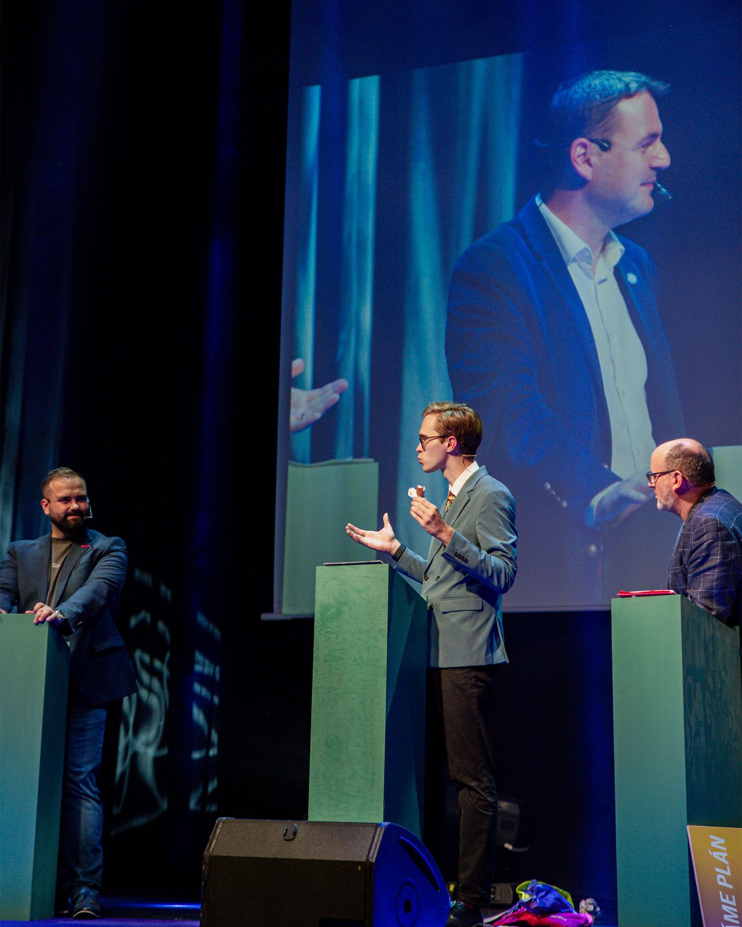 Three men on conference stage with large screen projection showing speaker in background.