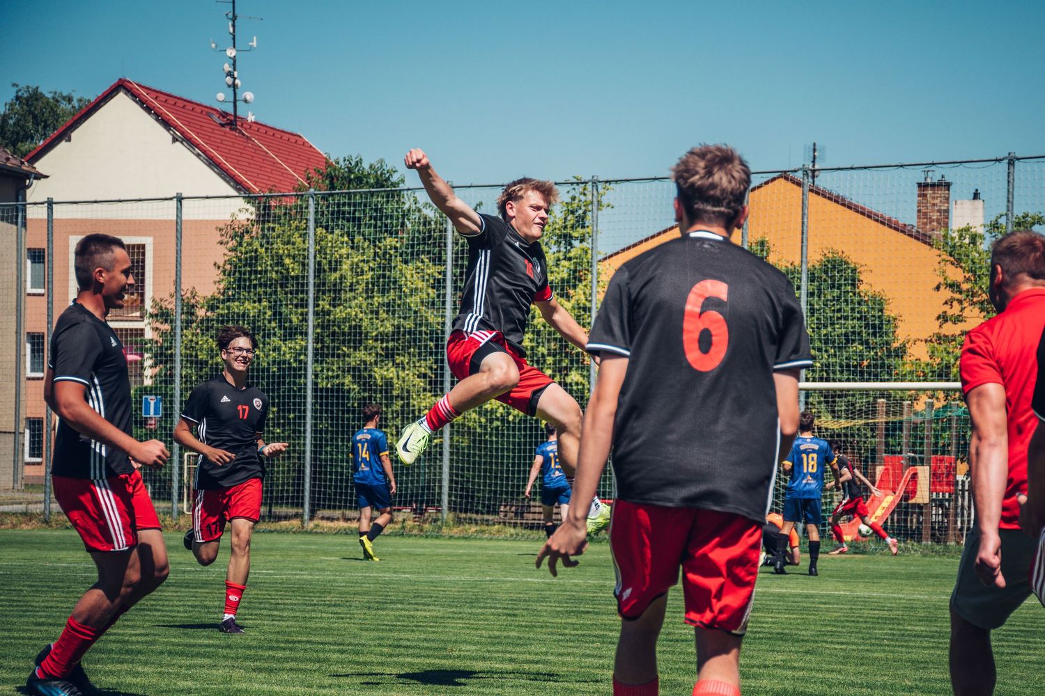 Young soccer players in black and red uniforms celebrate on a green field during a sunny outdoor match.