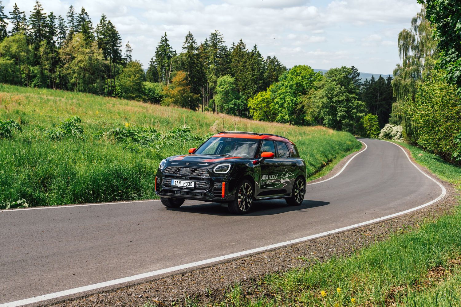 Orange MINI Cooper JCW driving on a curved road through a lush green countryside landscape.