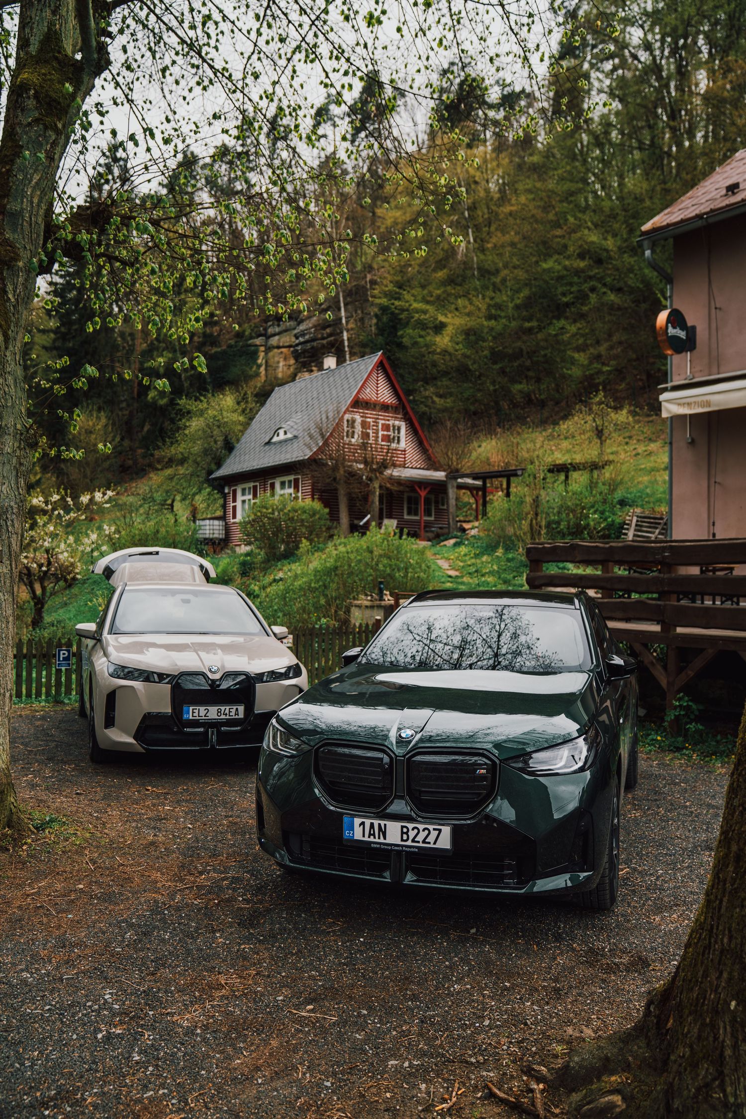 Two BMW electric SUVs, iX and XM, parked on gravel in front of a charming woodland cottage.