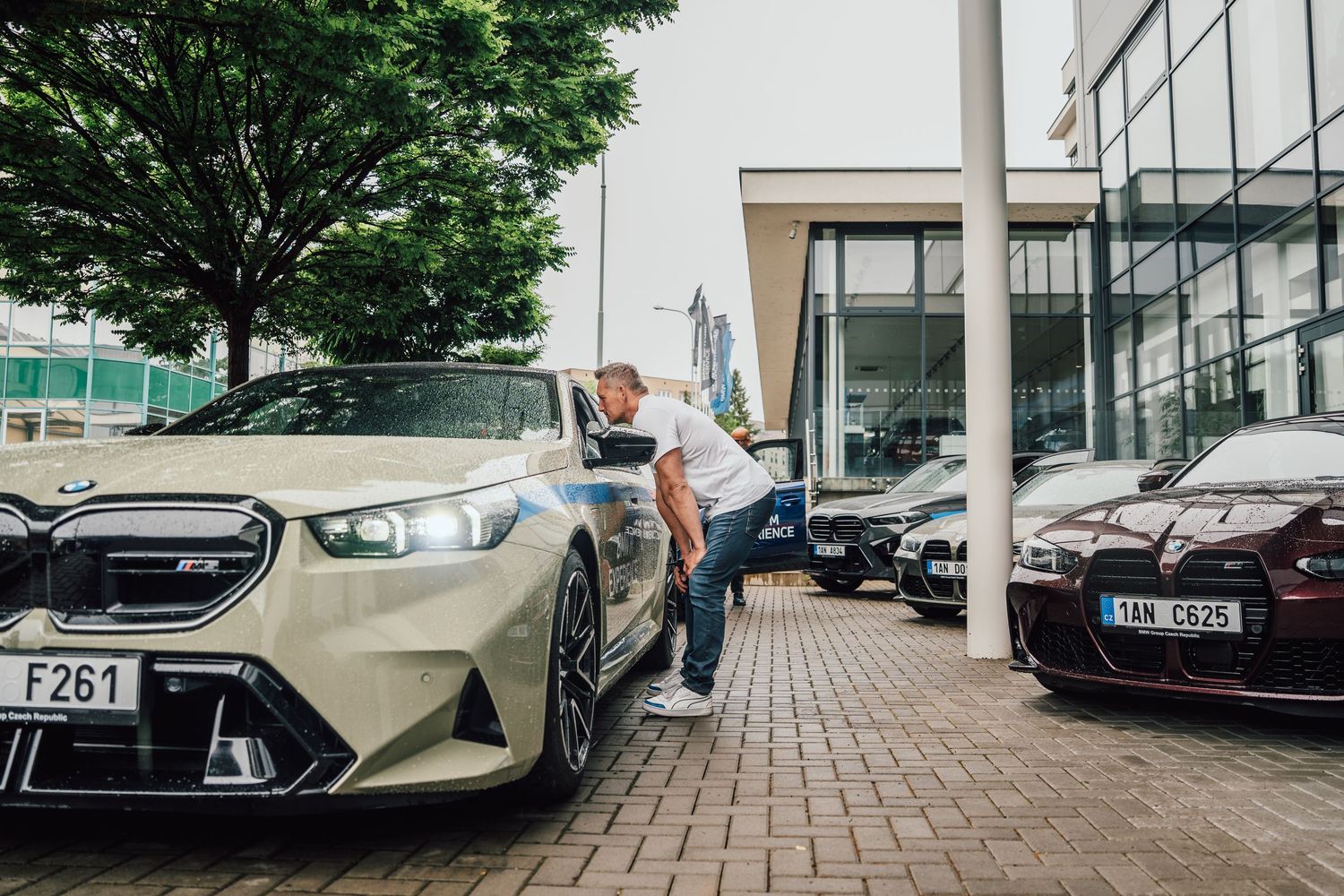 Man inspecting a gold BMW M2 at a BMW dealership with multiple luxury cars parked outside.