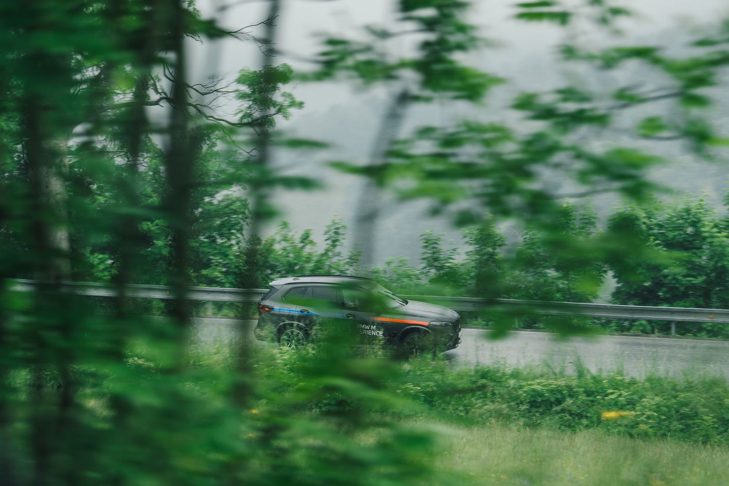 A dark-colored sports car driving on a wet road surrounded by lush green trees in rainy conditions.