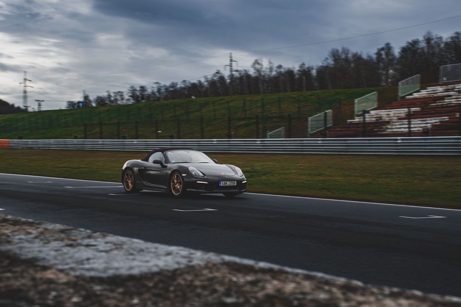 Dark grey Porsche Boxster GTS driving on a wet race circuit under overcast skies.