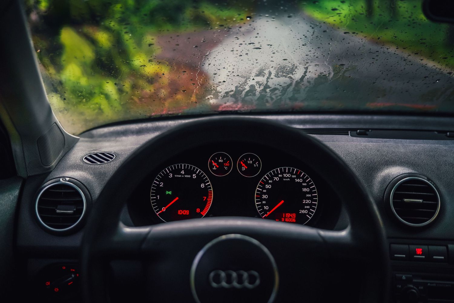 Audi dashboard with illuminated gauges viewed through rain-covered windshield on a wet road.