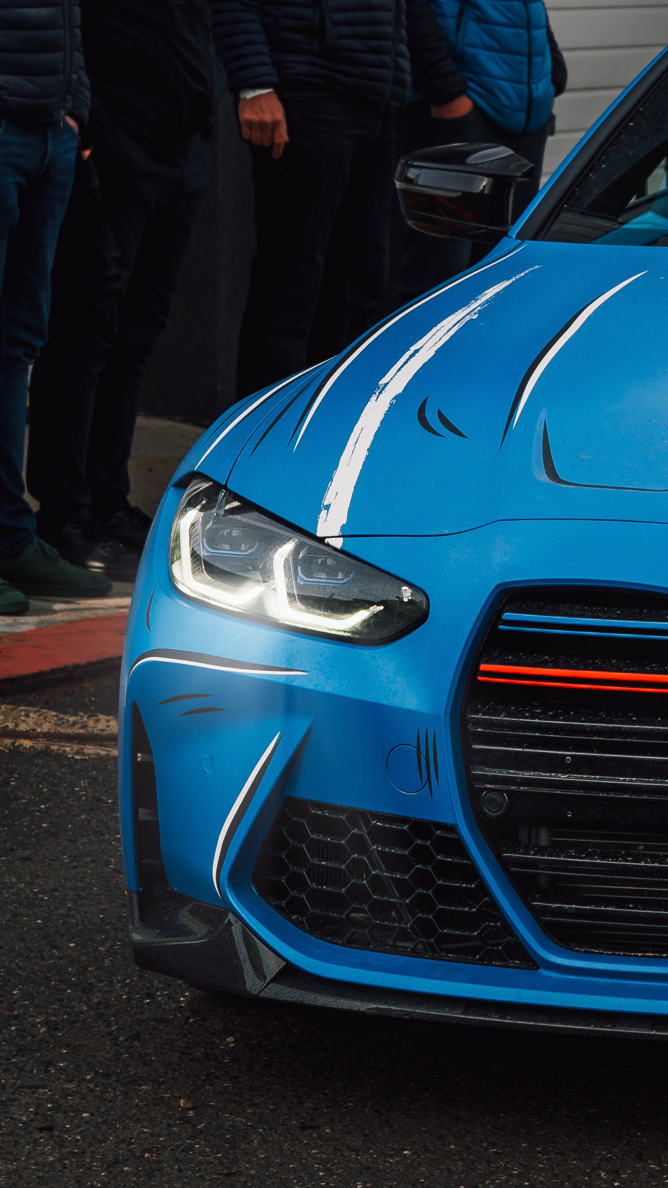 Blue BMW M2 Competition front view with aggressive styling, LED headlights, and carbon fiber splitter at a motorsport event.