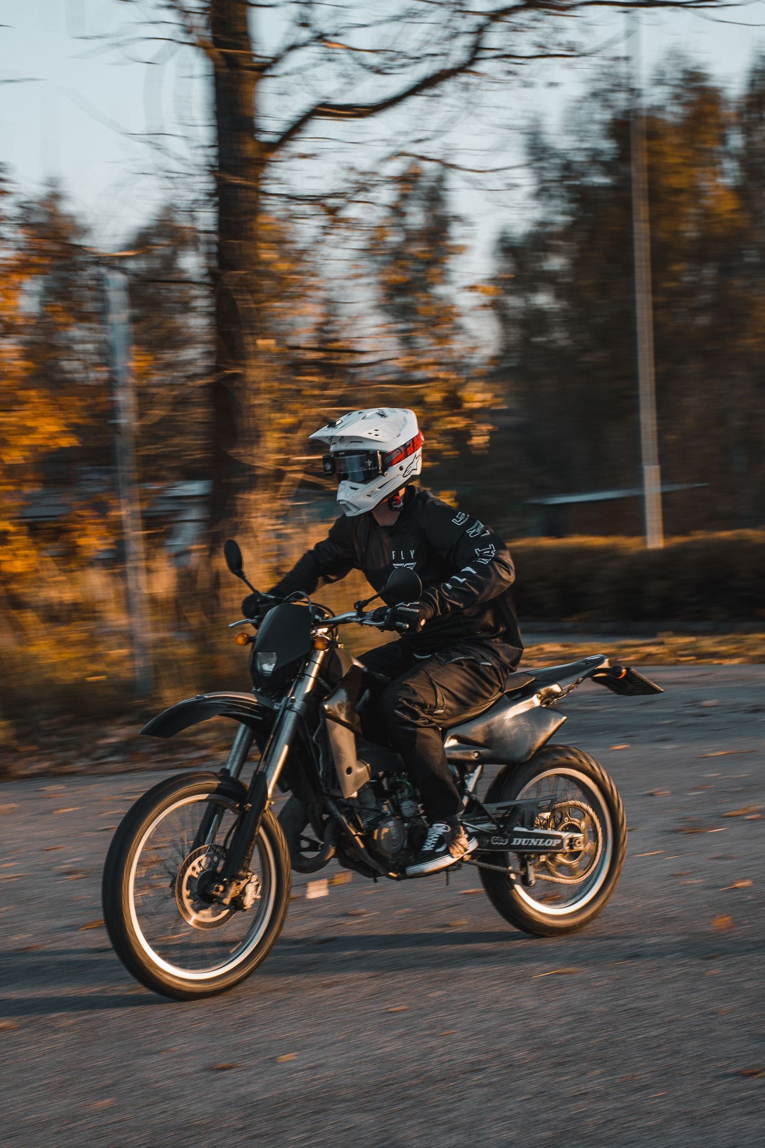 Rider in white helmet on black supermoto motorcycle speeding on road with autumn trees backdrop.