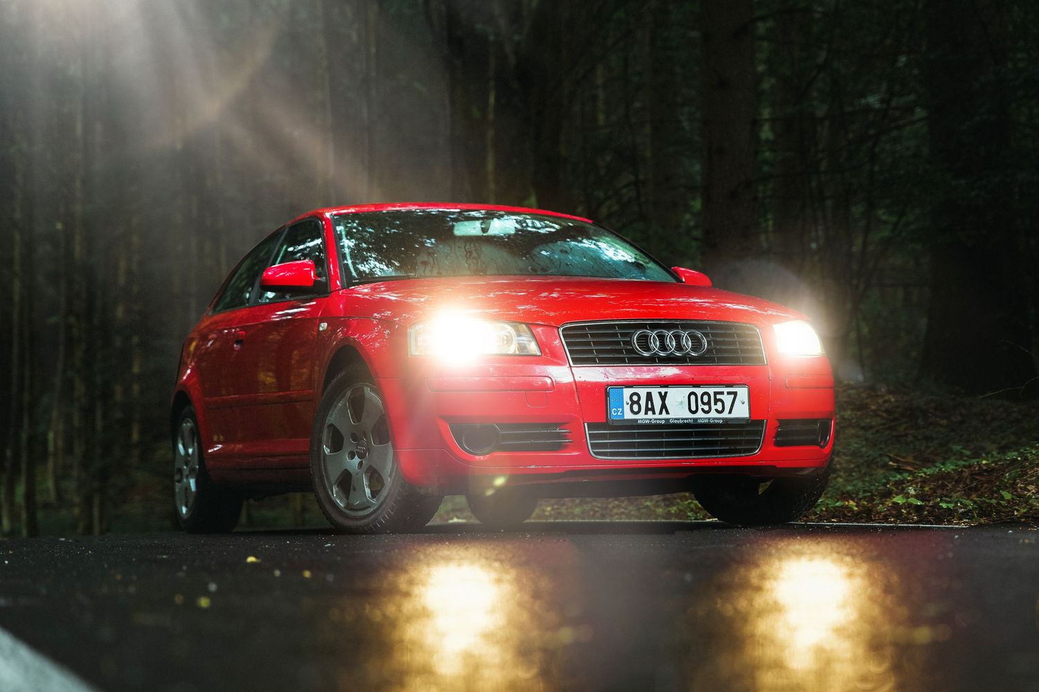 Red Audi TT with headlights on driving on a wet forest road in rainy conditions.