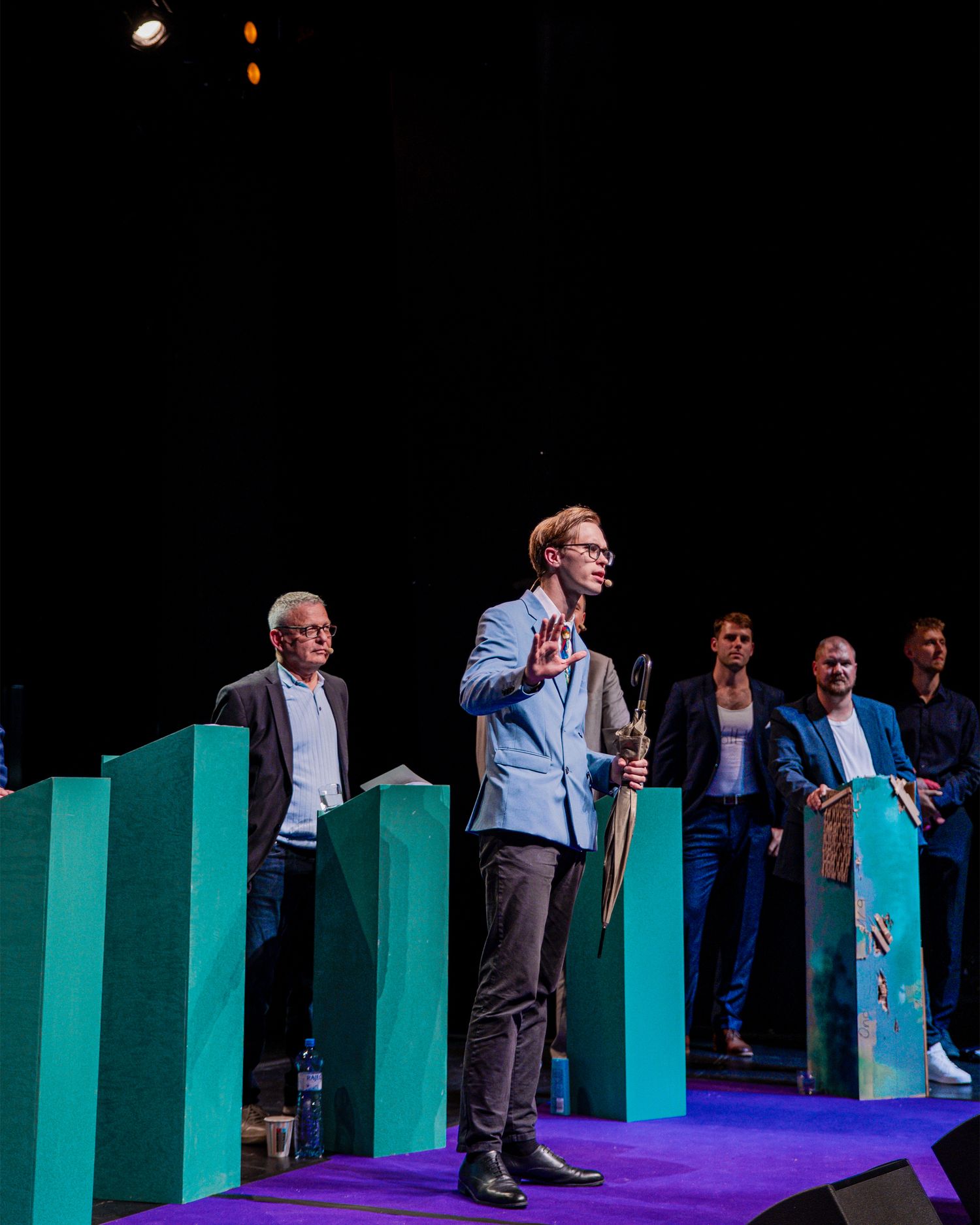 Group of men in suits standing at teal podiums on a stage during a formal conference or award ceremony.