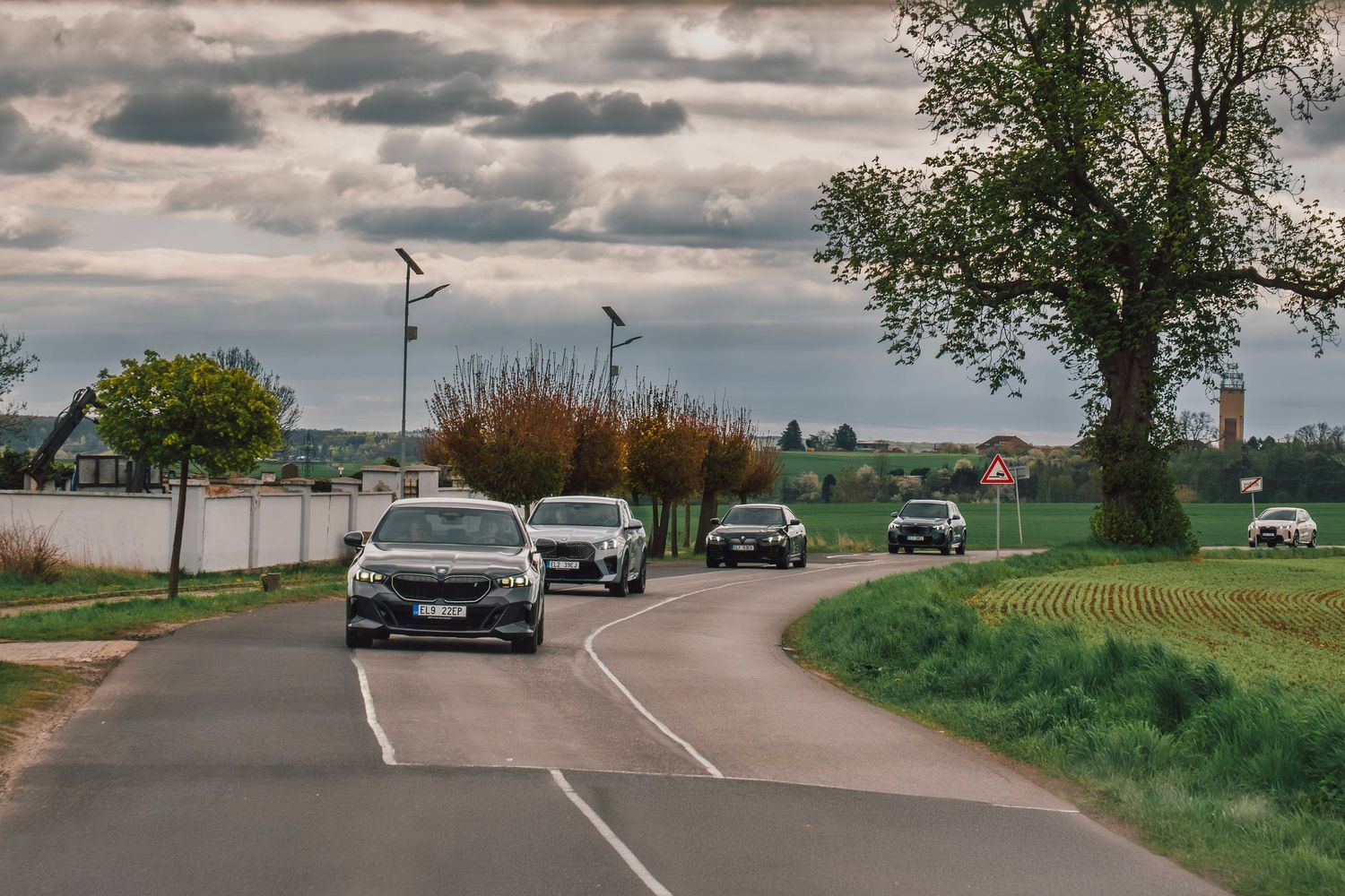 A convoy of BMW electric vehicles driving along a rural road under dramatic cloudy skies with green fields.