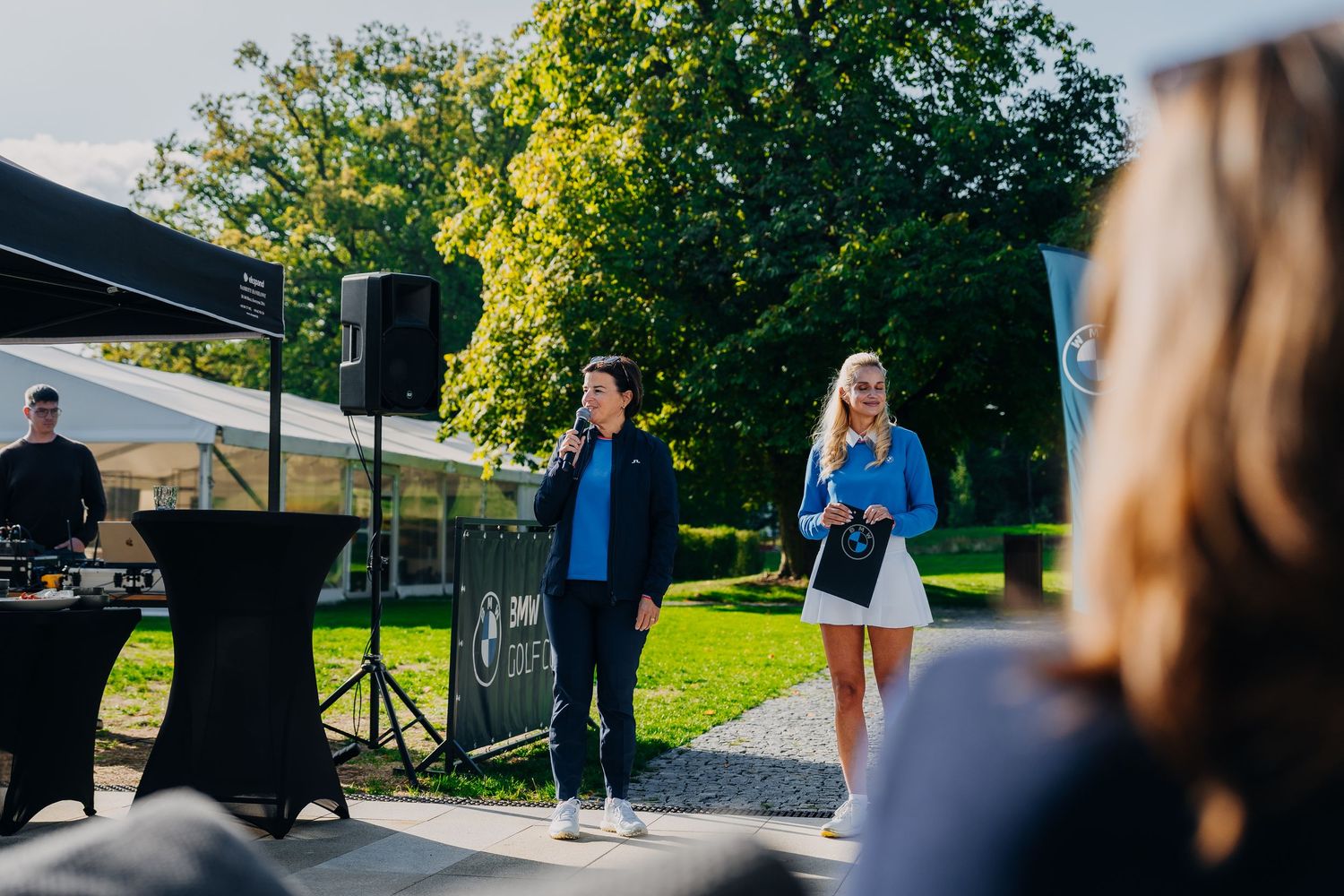 Two women presenting outdoors at a corporate event with speaker and greenery in background.