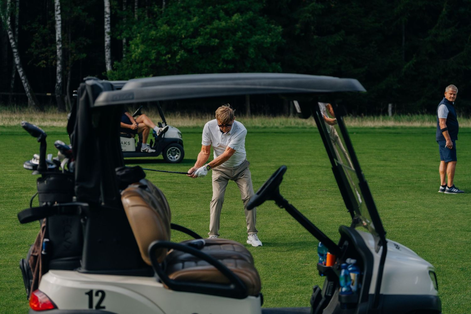 Golfers playing on a lush green golf course with golf carts nearby and trees in the background.