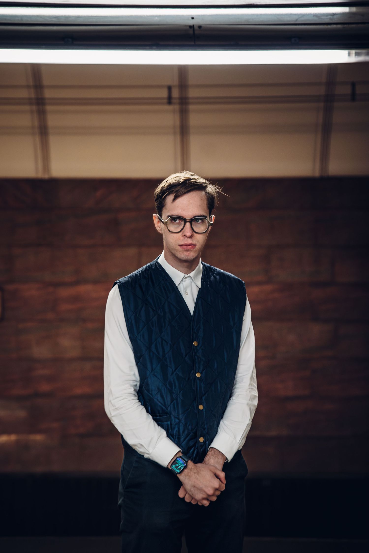 Young man in glasses and blue vest standing confidently in front of a brick wall.