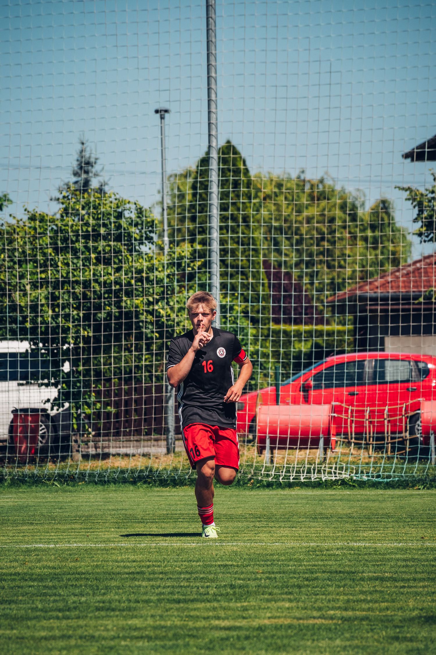 Young soccer player in black and red uniform walks on green field near goal net.
