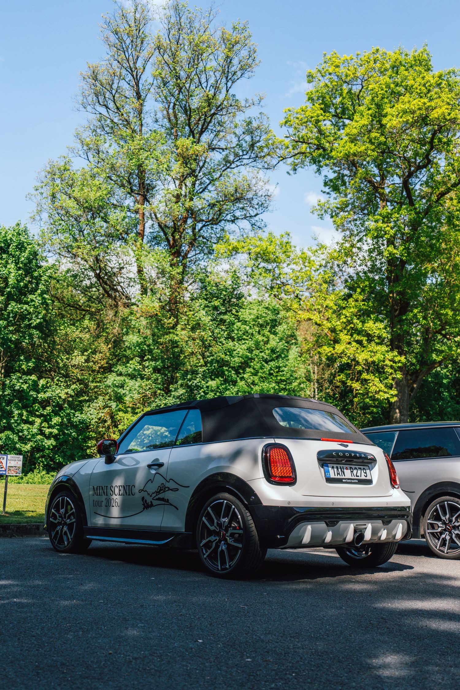 White MINI Cooper convertible with custom graphics parked outdoors under a bright blue sky with green trees.