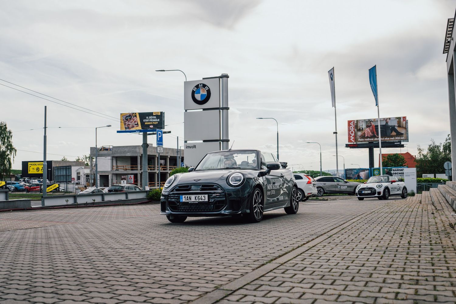 Black MINI Cooper convertible parked at a BMW dealership under an overcast sky.