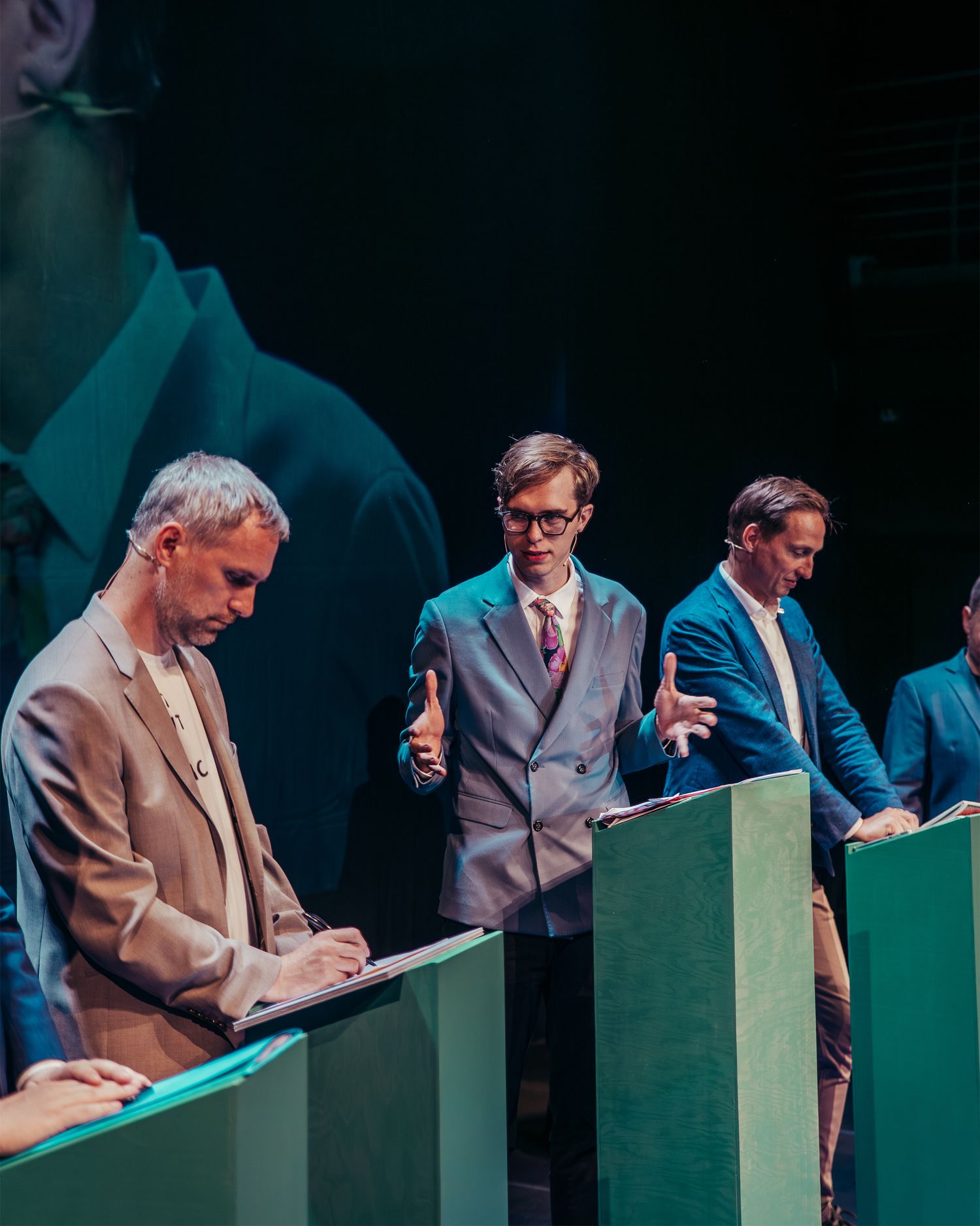 Three men stand at green debate podiums on a dramatic stage with projection.