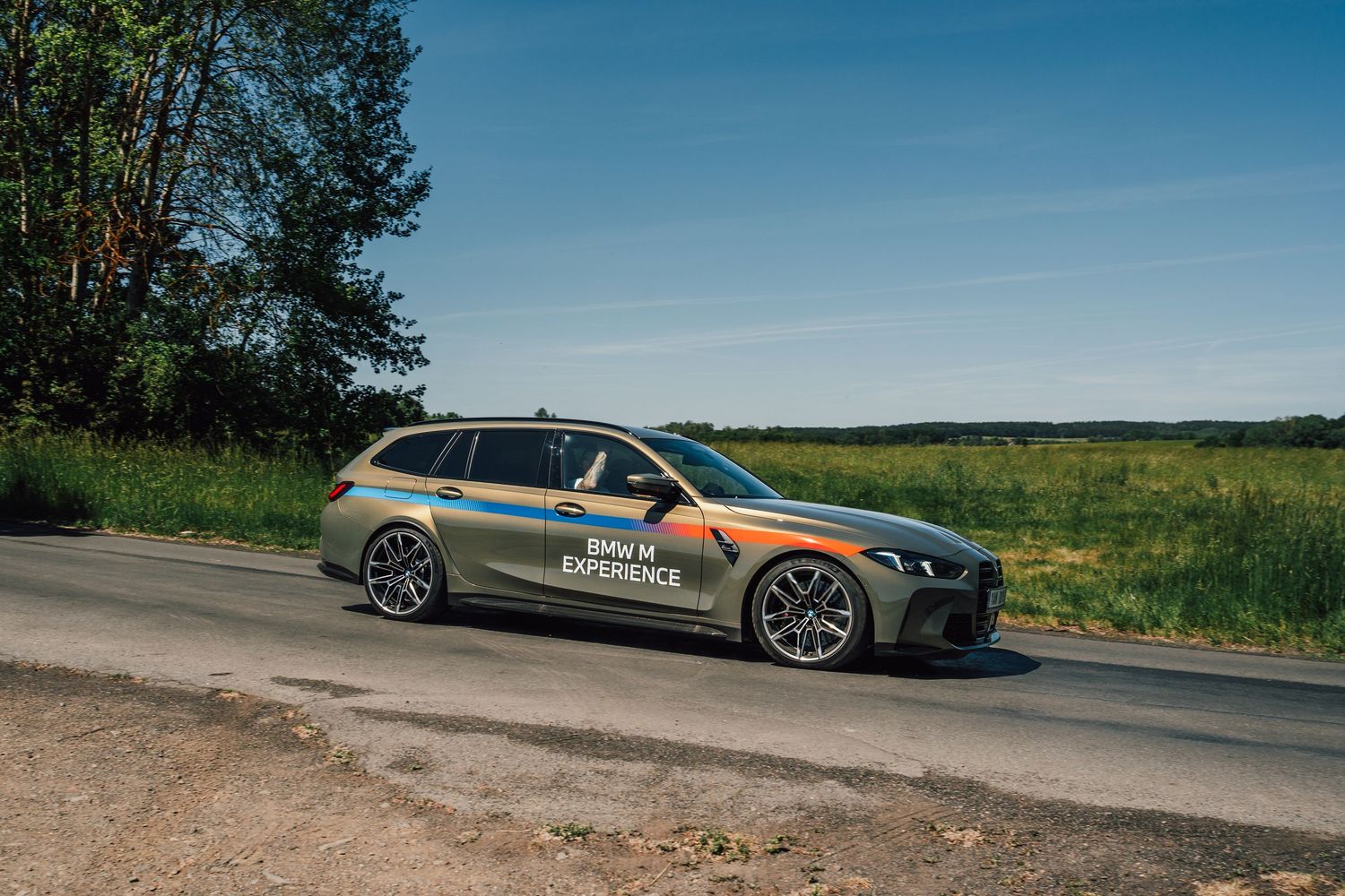 BMW M3 Touring with BMW M Experience livery parked on a rural road beside green fields.