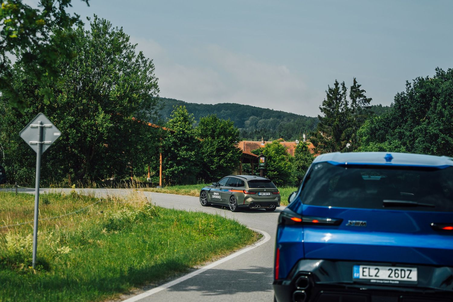 A blue BMW iX and grey BMW M3 Touring navigating a winding rural road in scenic countryside.