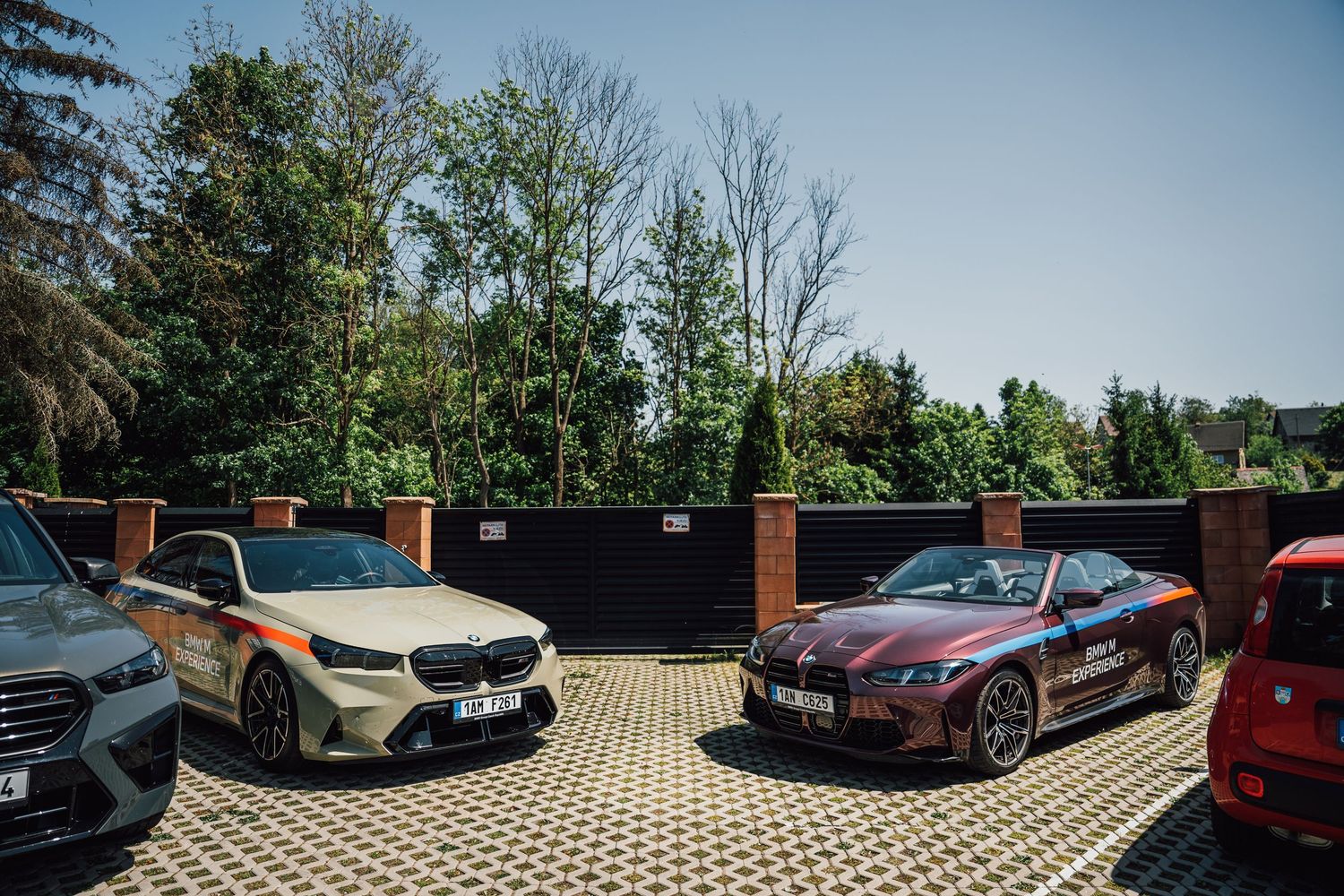 Multiple BMW M Experience cars including a convertible parked in a courtyard surrounded by trees.