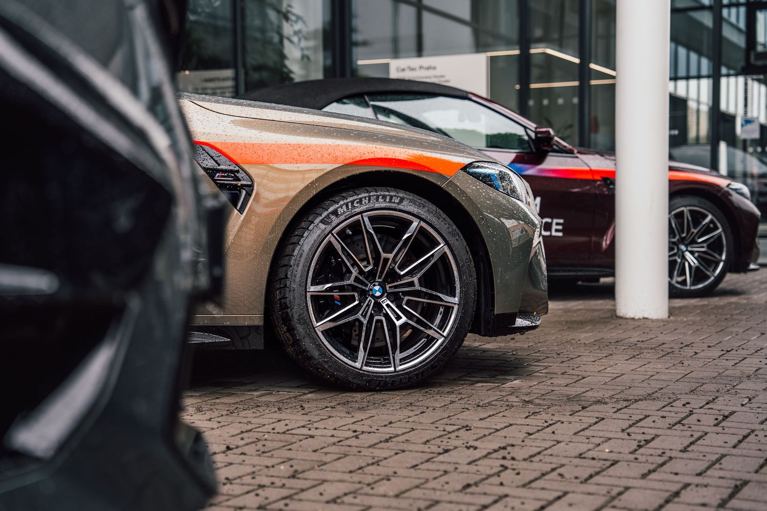 Row of BMW M cars parked at a dealership, featuring a striking two-tone gold and green wrap on the center vehicle.