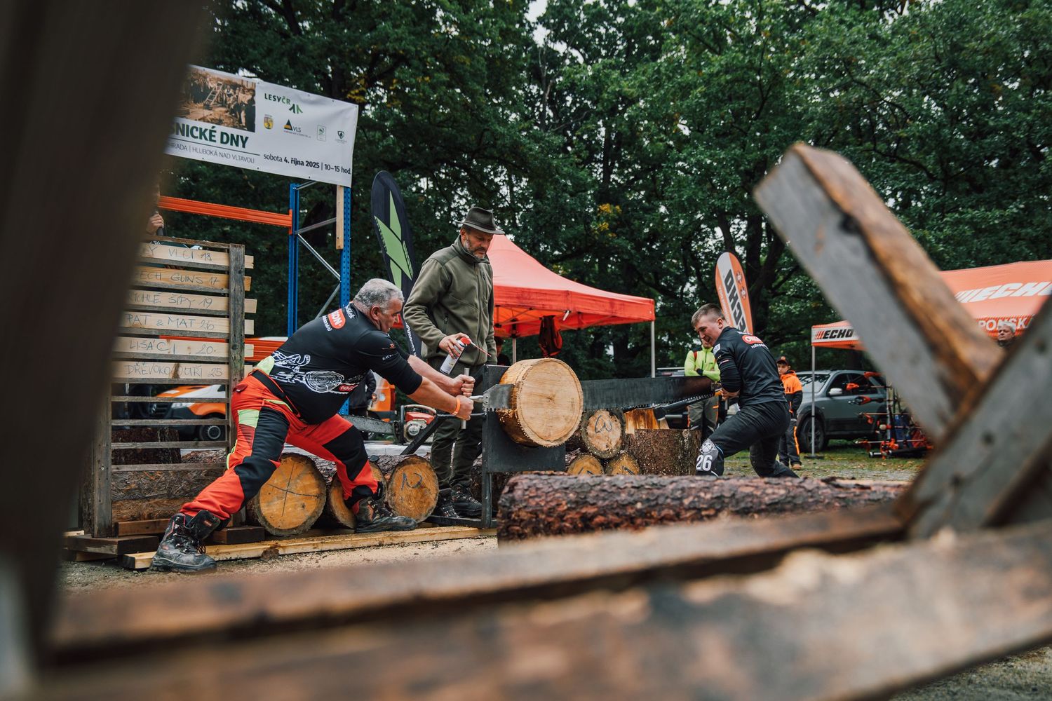 Two competitors use chainsaw to cut logs together at outdoor lumberjack sports competition event.