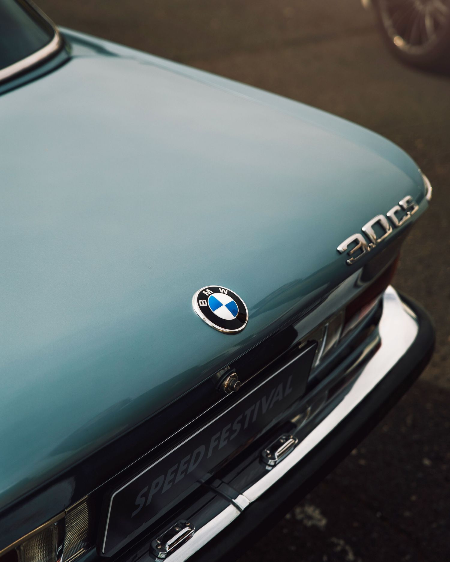 Close-up of a vintage light blue BMW 2002 rear badge and Speed Festival license plate in moody lighting.