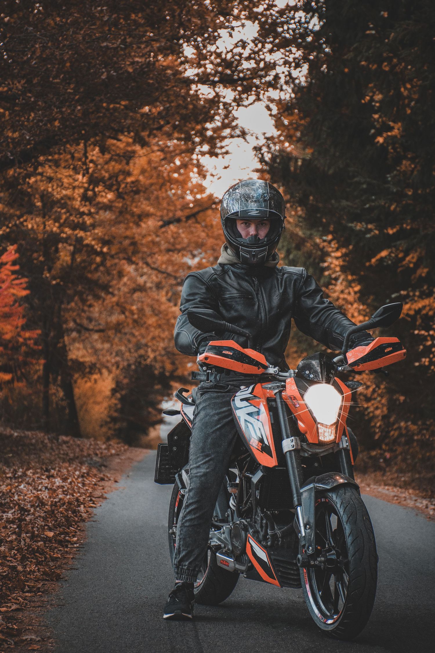 Motorcyclist in black gear sitting on an orange and black KTM Duke on an autumn forest road.