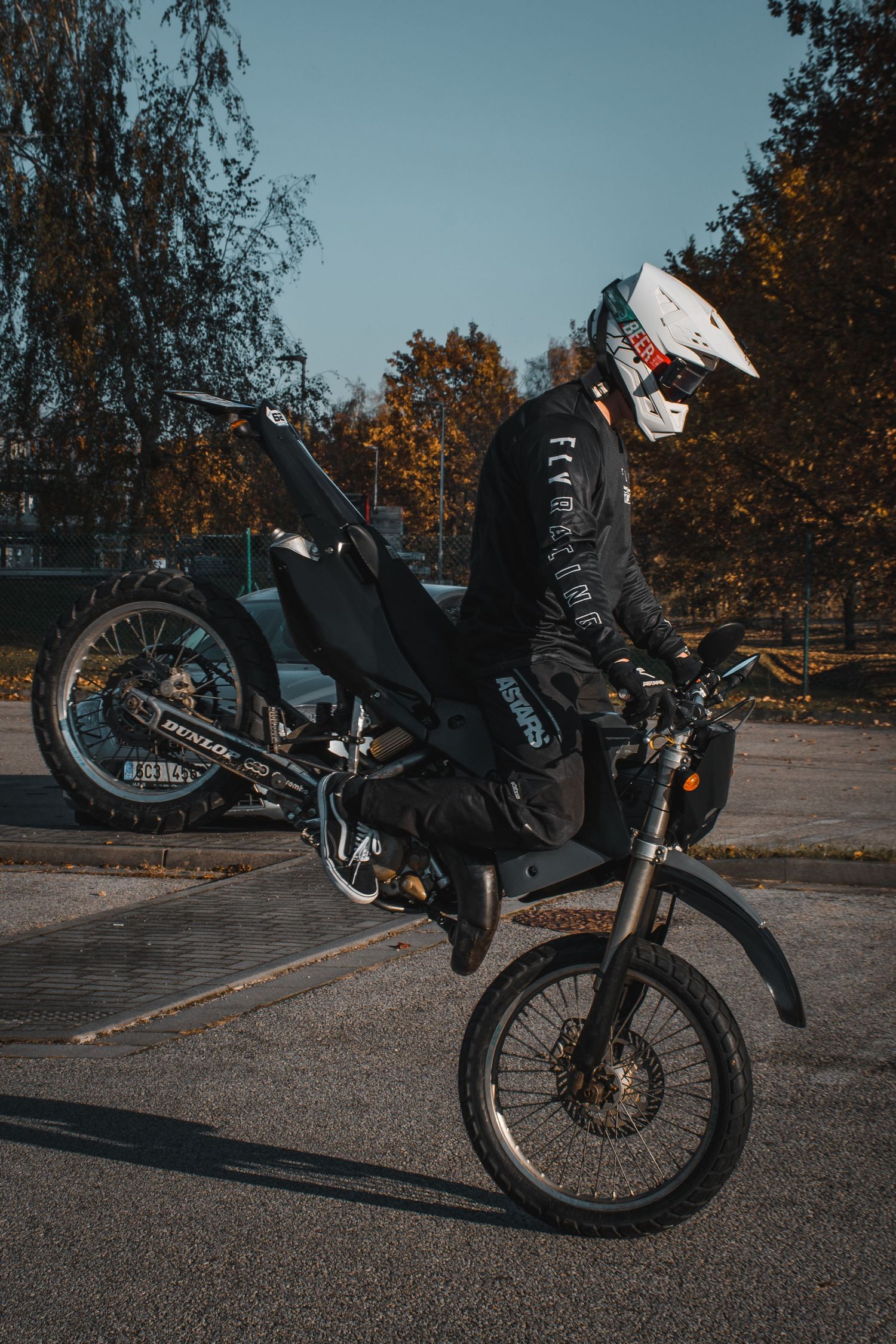 Stunt rider performing wheelie on black dirt bike in parking lot with autumn trees in background.