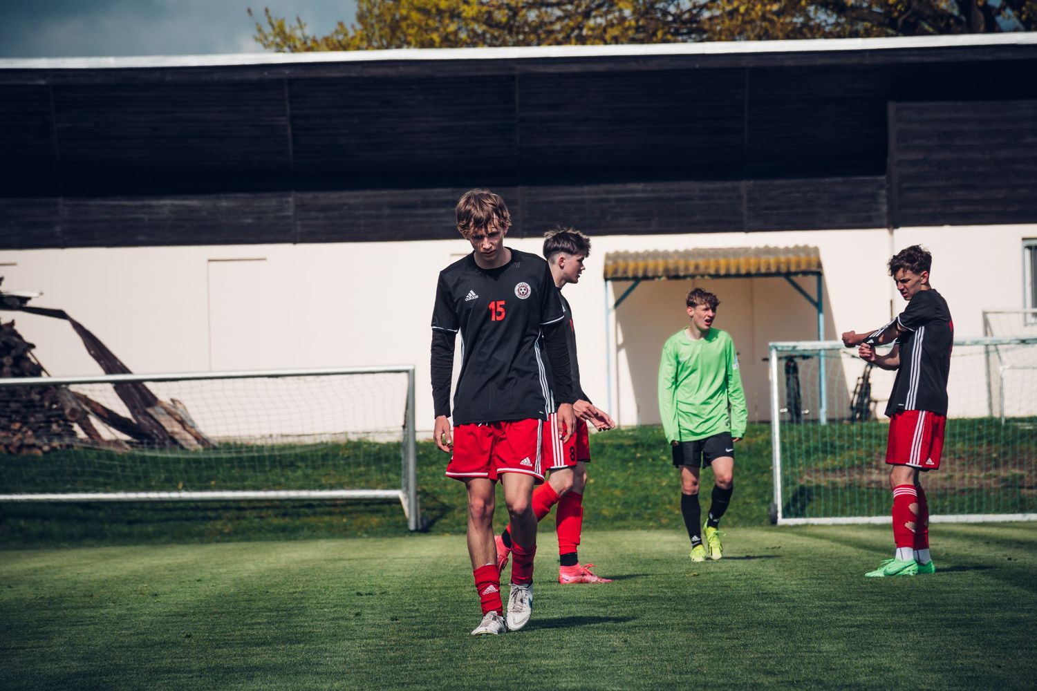 Young soccer players in black and red uniforms walk near the goal post on a green field during a match.
