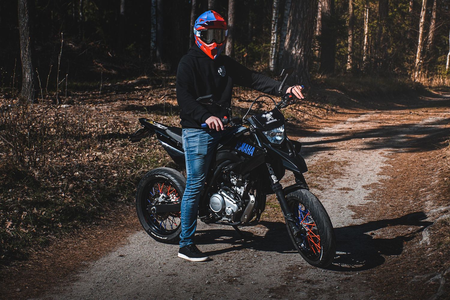 Rider in blue jeans and red helmet standing beside a black supermoto on a forest path.