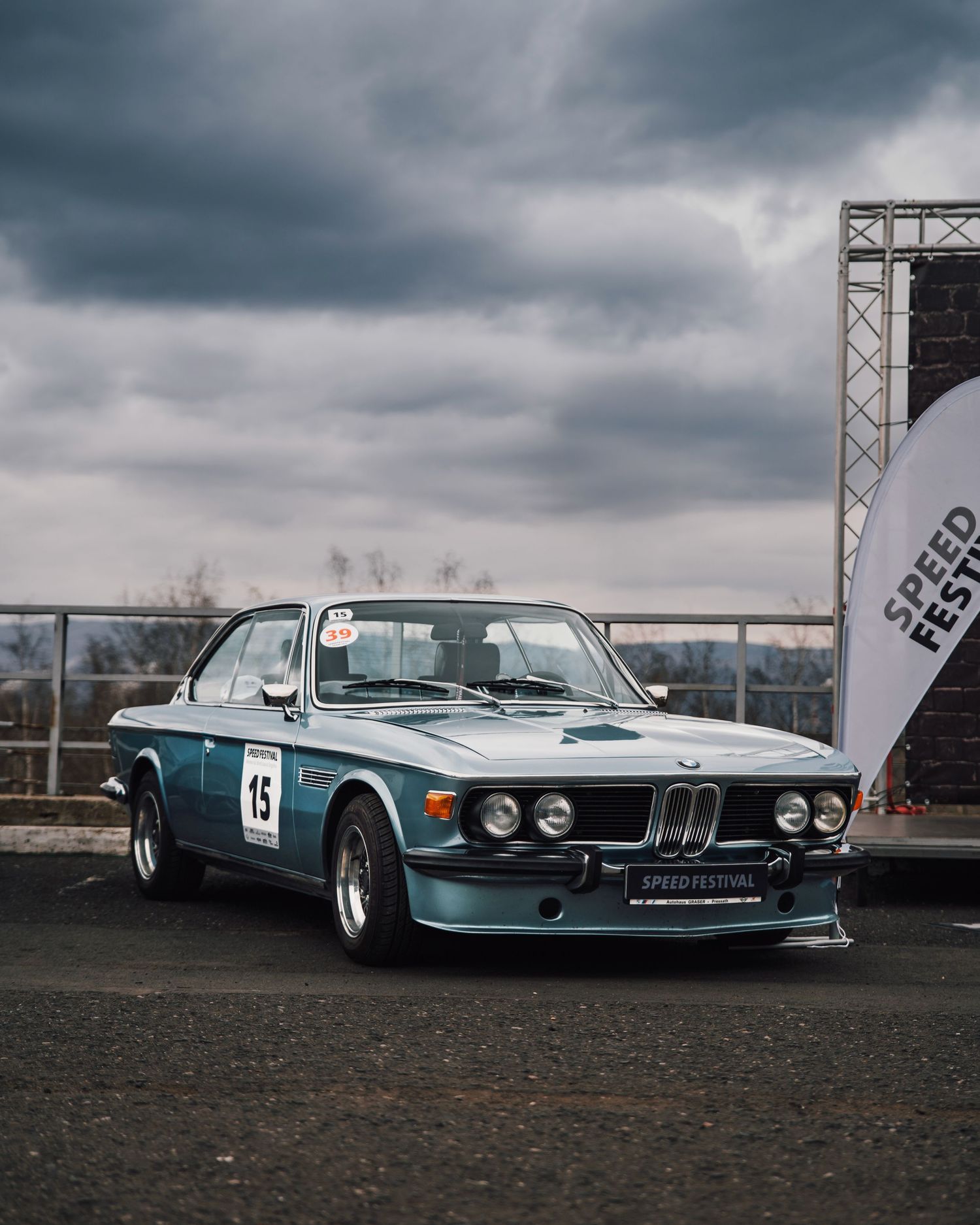 Vintage blue BMW 3.0 CSL race car number 15 parked at Speed Festival event under dramatic cloudy sky.
