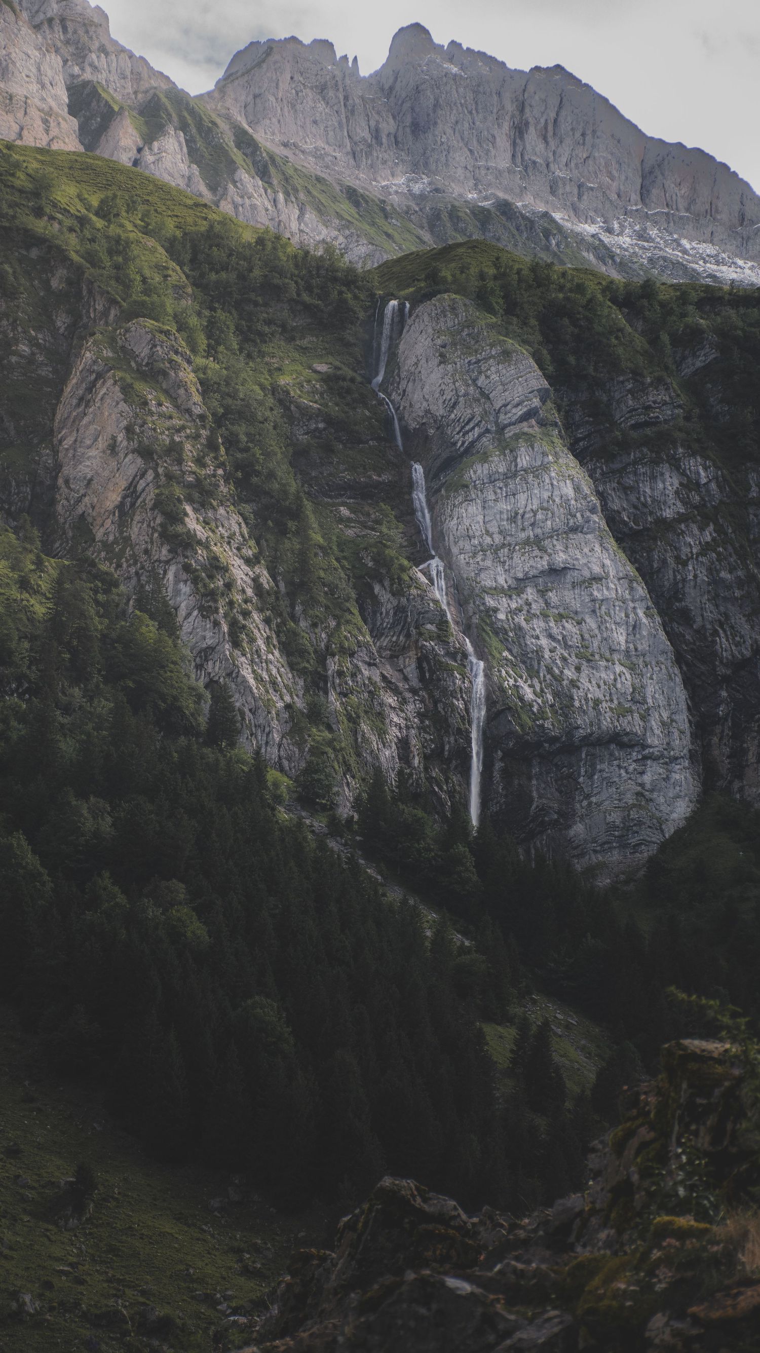 A dramatic waterfall cascades down a steep rocky cliff face surrounded by lush green forest in the mountains.
