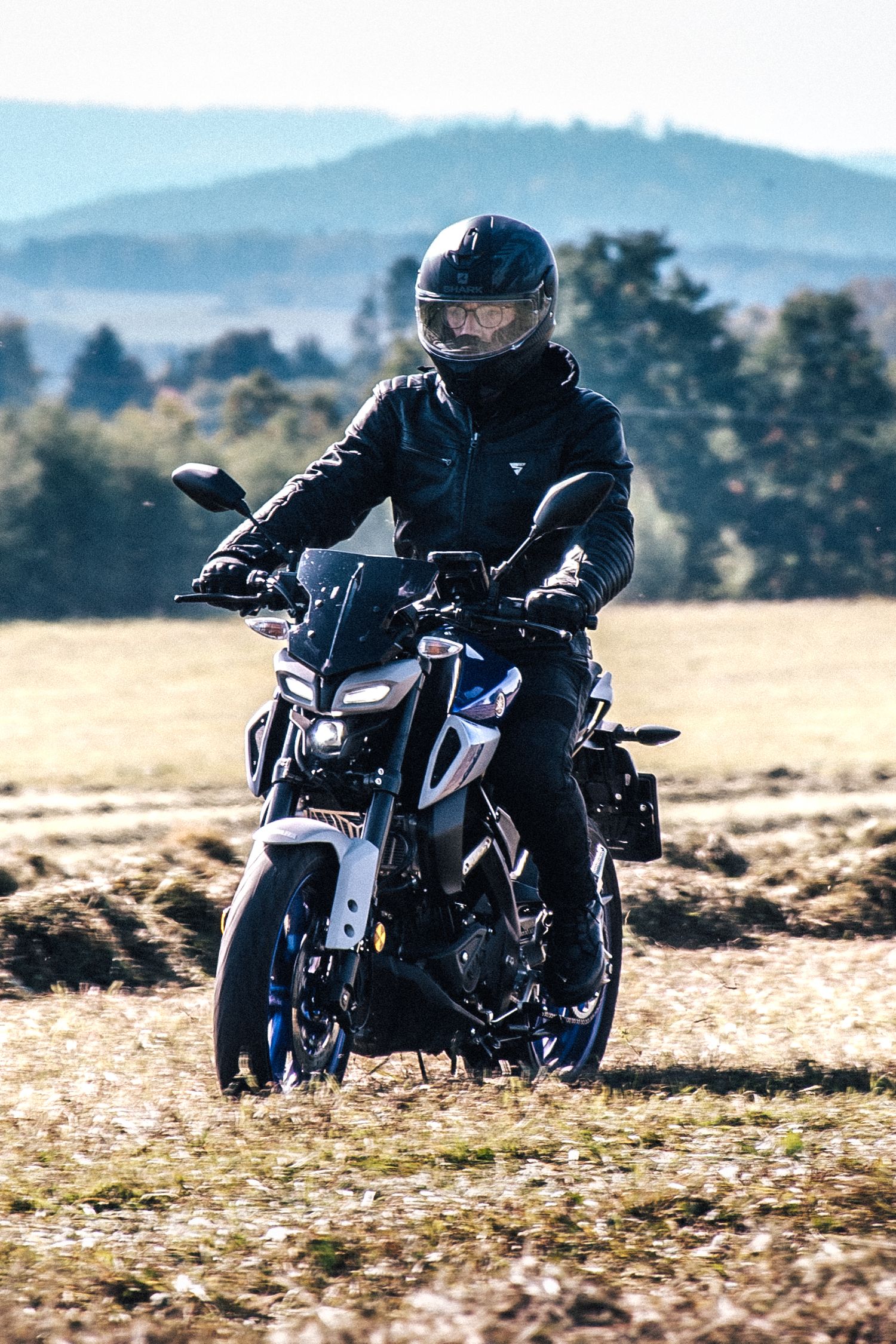 Rider in full black motorcycle gear on a blue naked bike parked in an open field with mountain backdrop.
