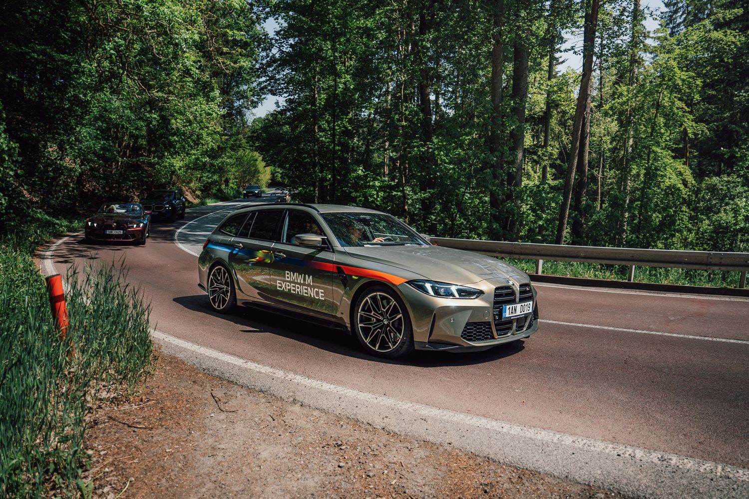 BMW M3 Touring with M Experience livery driving on a forest road flanked by tall green trees.