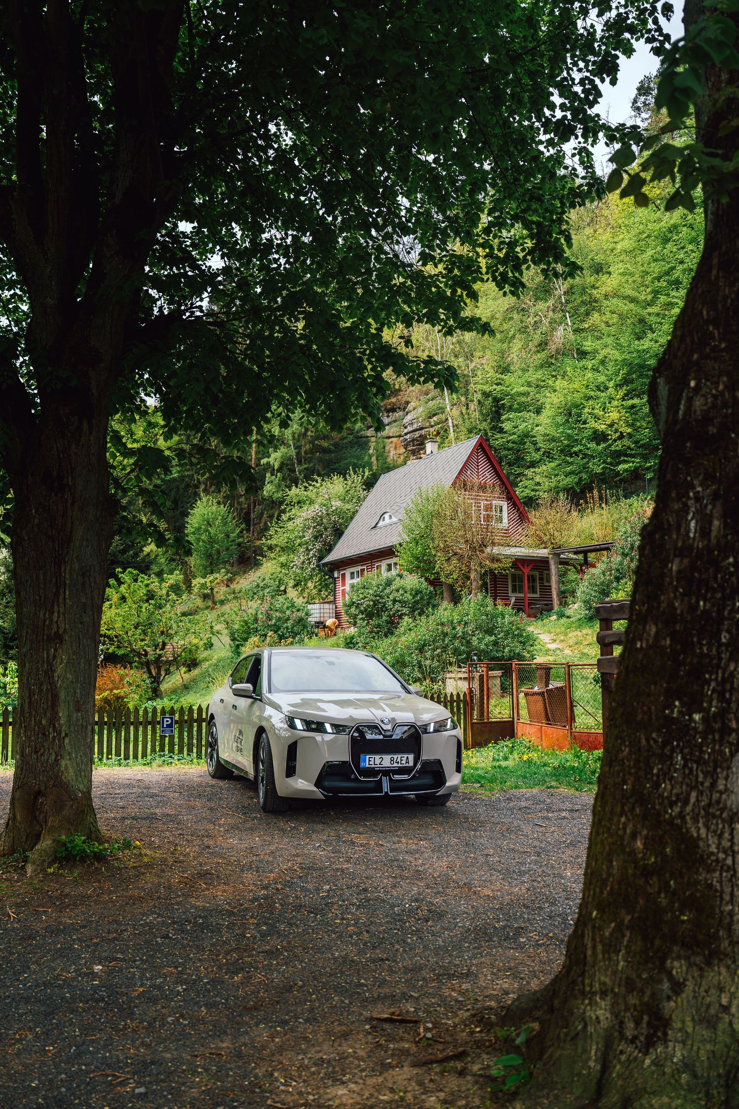 Silver BMW electric SUV parked on a gravel driveway framed by tall trees with a rustic cottage behind.