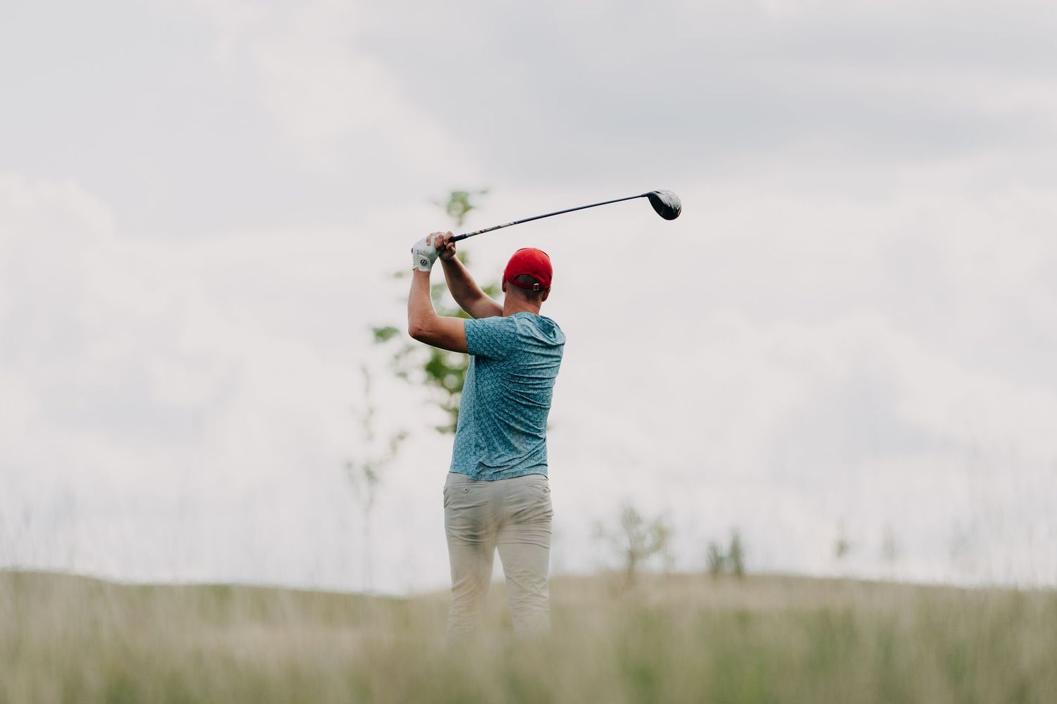 Golfer in blue shirt and red cap swings a driver on a lush green golf course under cloudy skies.
