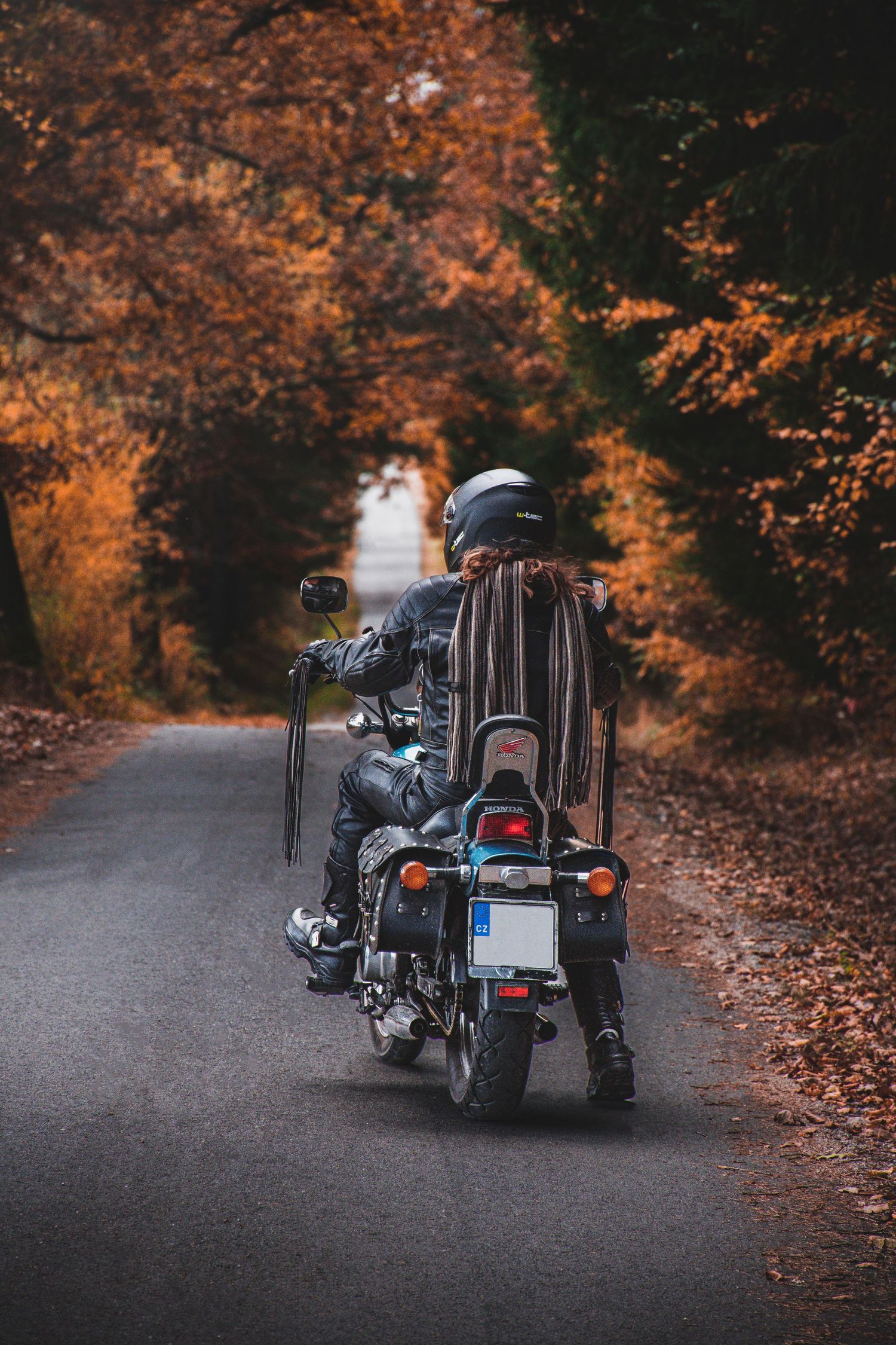 Rear view of a motorcyclist with long hair riding a classic cruiser motorcycle on an autumn forest road.