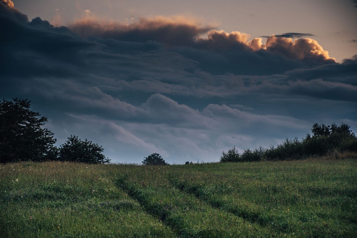 Dirt tracks through green meadow under dramatic stormy sky with glowing orange-lit rolling clouds at dusk.