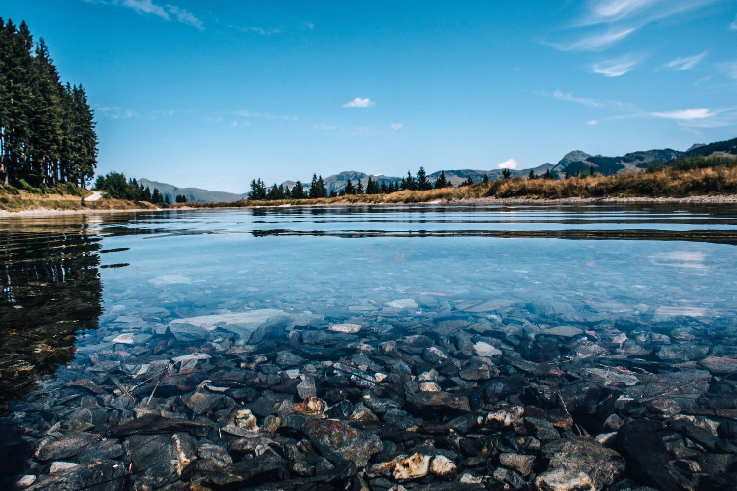 Crystal clear shallow lake with rocky bottom reflecting blue sky and surrounding pine trees.