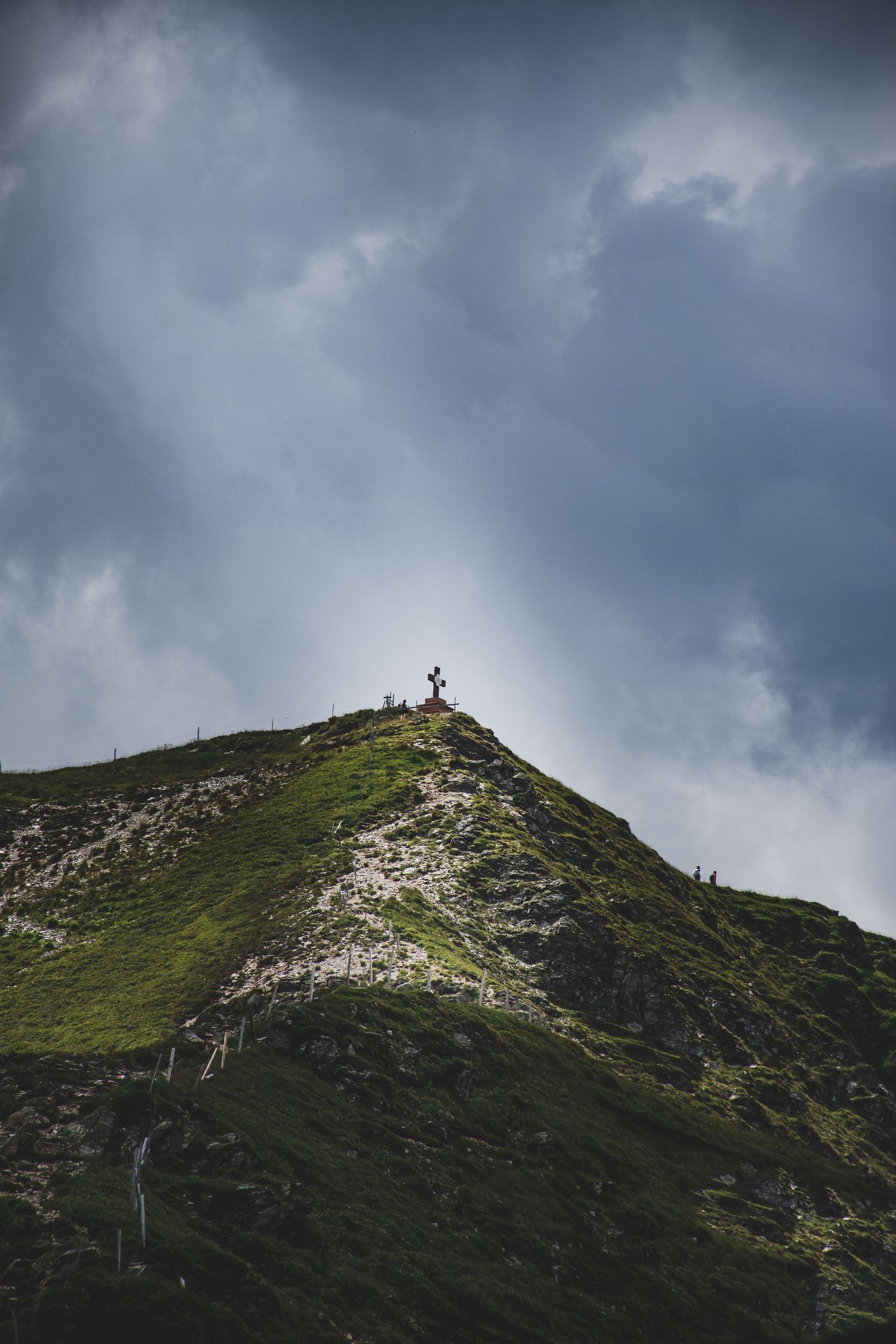 A green mountain summit with a wooden cross silhouetted against a dramatic stormy sky with dark clouds.