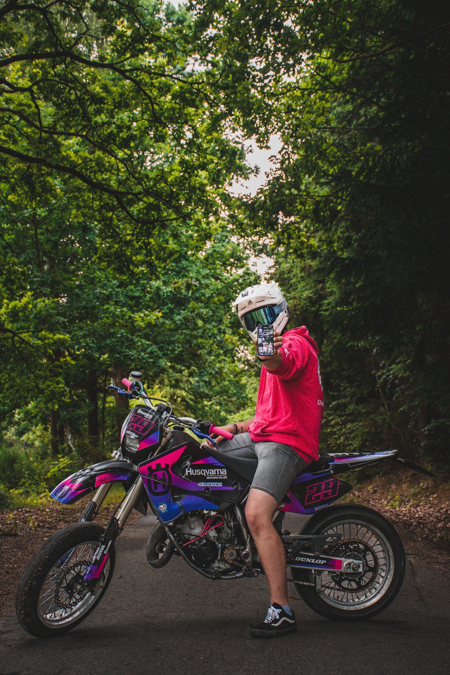 Motocross rider in red hoodie posing with Husqvarna supermoto bike on a forest road.
