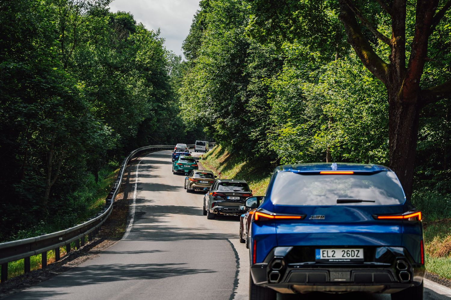 A convoy of BMW M cars led by a blue BMW iX driving along a tree-lined country road.