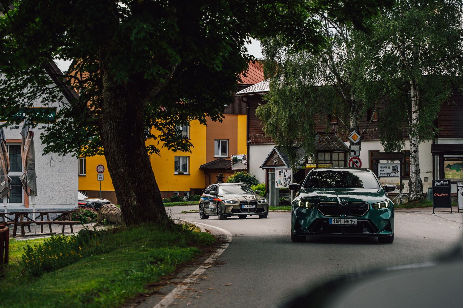 Green BMW M3 drives through a quaint European village with yellow buildings on a cloudy day.