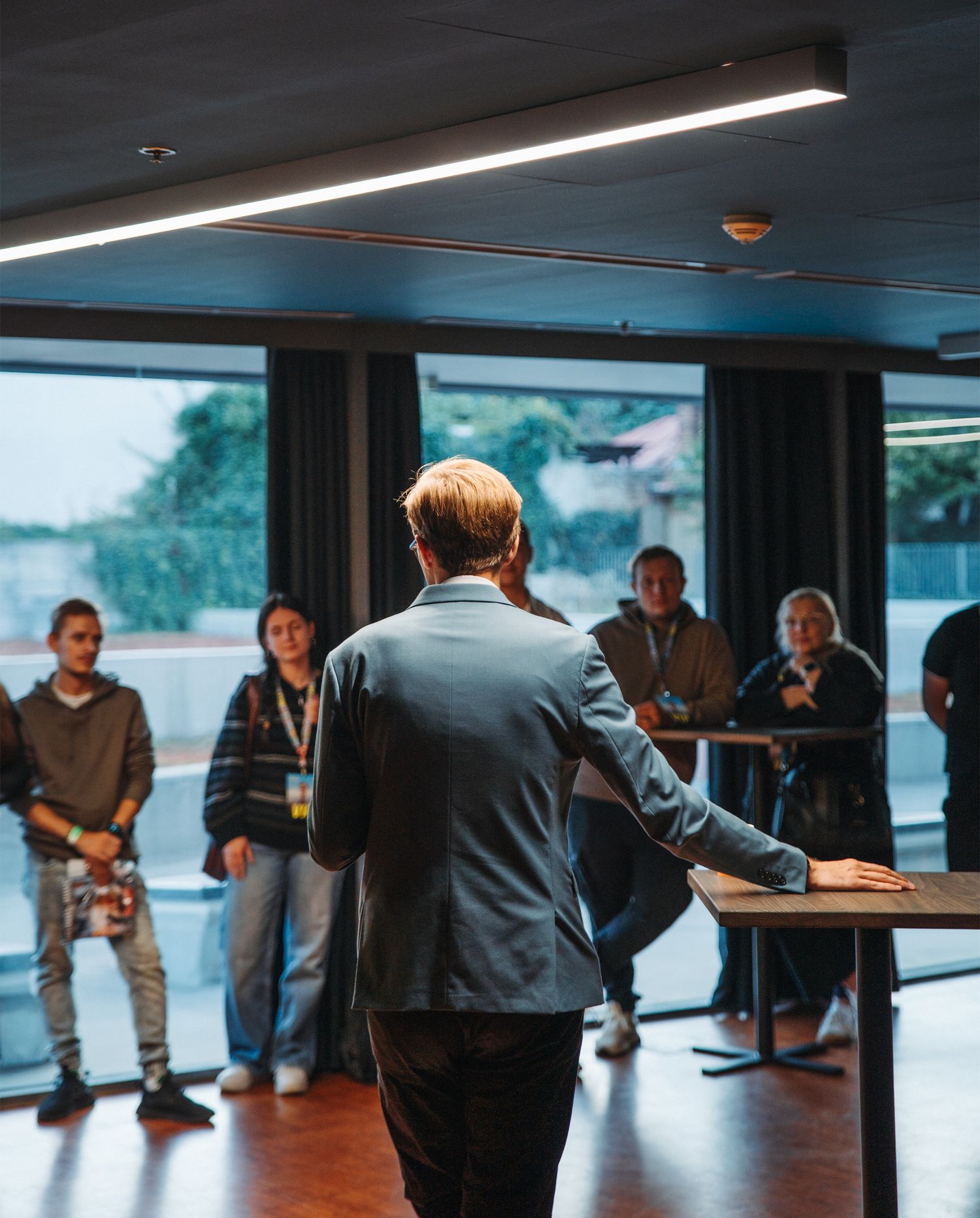 Speaker in gray blazer presenting to small group in modern room with large windows.