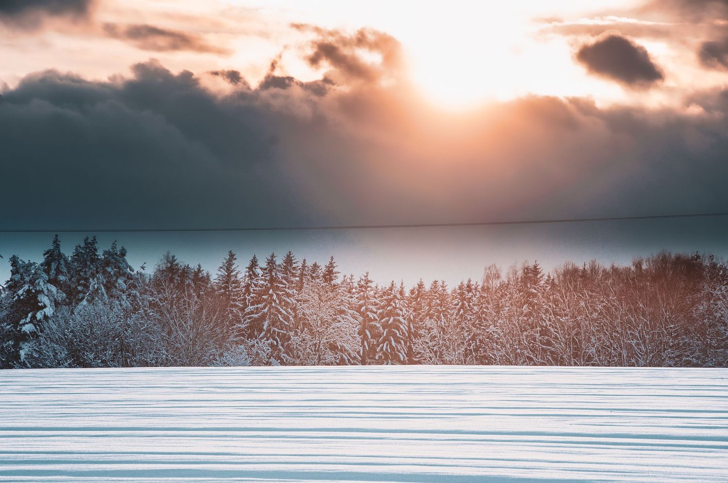 Golden sunset breaking through dramatic storm clouds over a snow-covered forest landscape in winter.