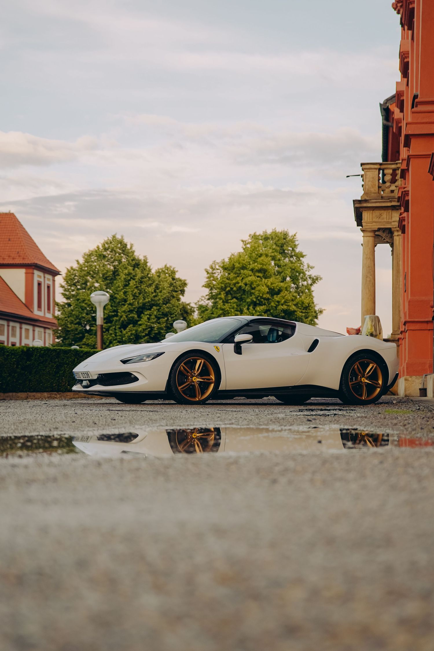 White Ferrari 296 GTB with gold wheels reflected in puddle outside historic European building.