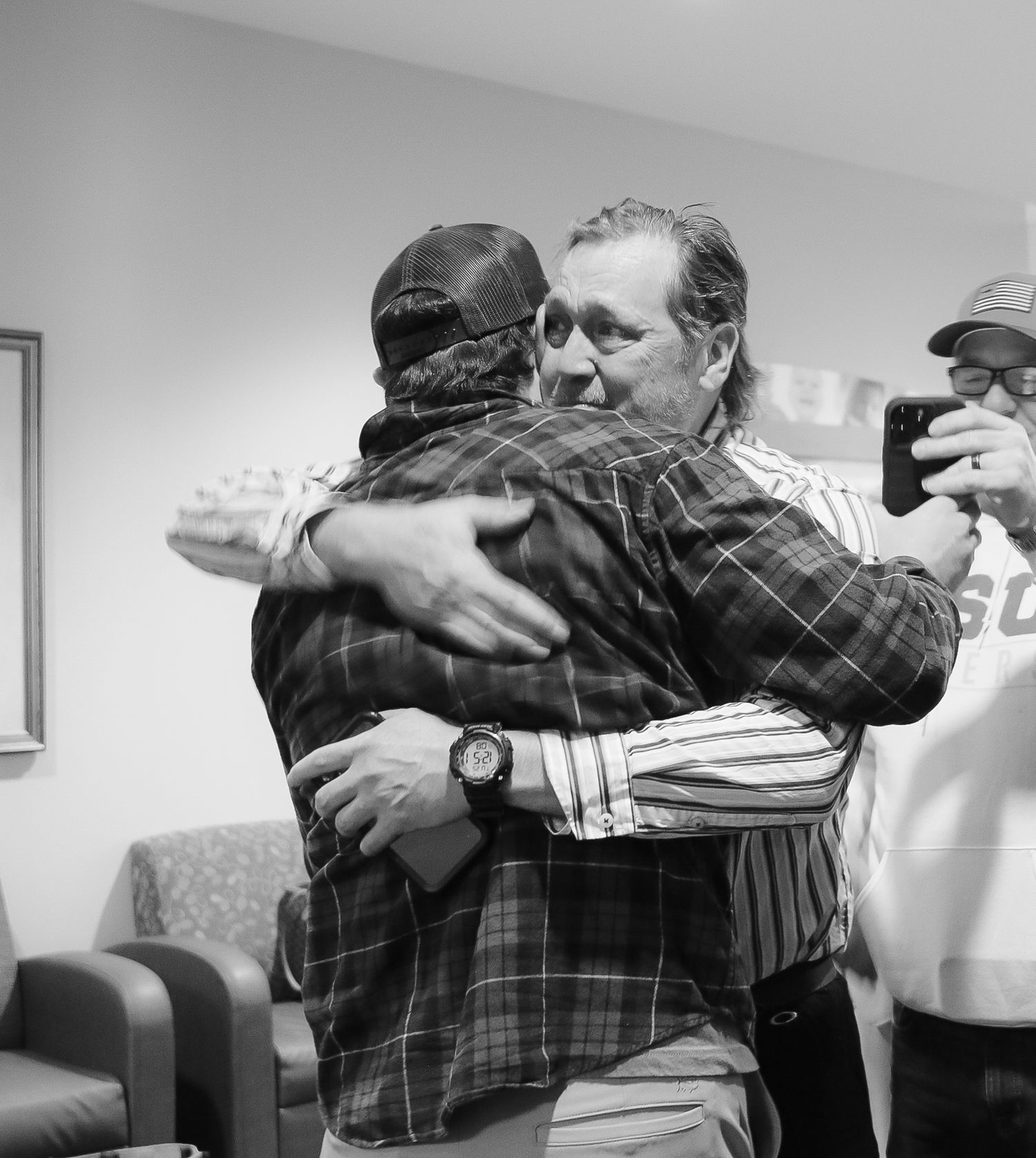 Two men share a warm embrace in an indoor setting while another person captures the moment on a smartphone.