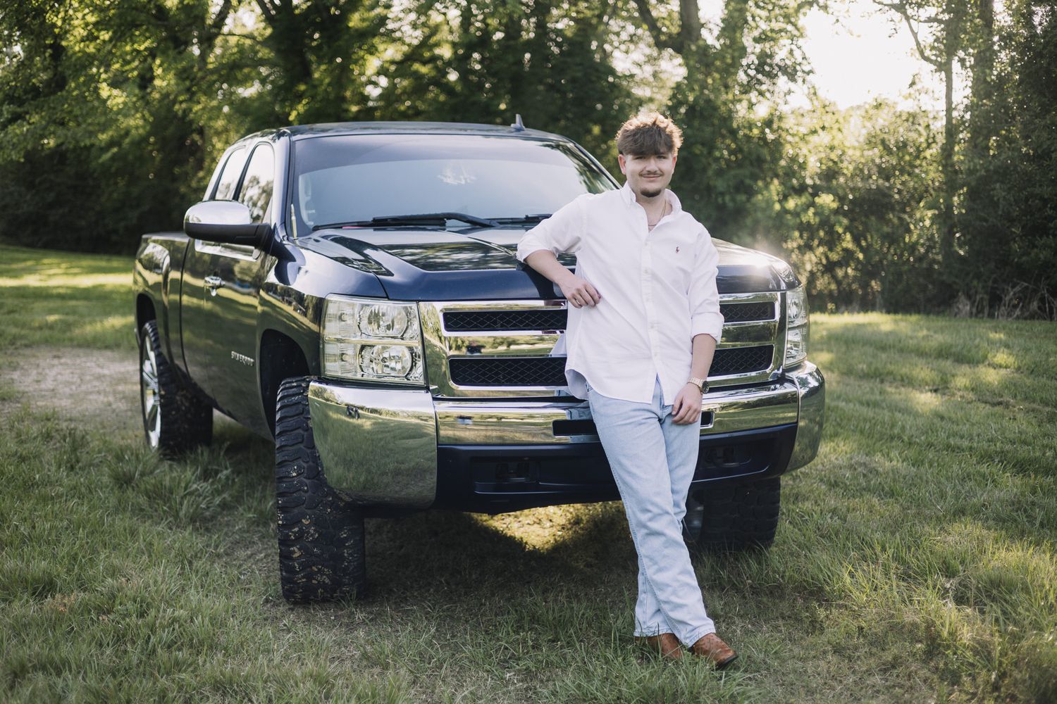 Man in white shirt and jeans leaning against a lifted black Chevrolet Silverado truck outdoors.